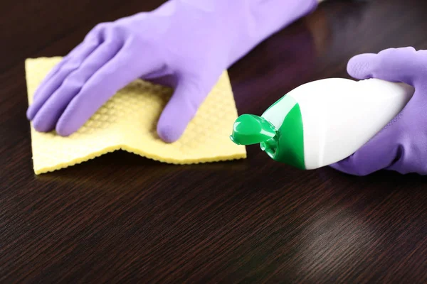 Hands in gloves with sponge and bottle of detergent cleaning kitchen table