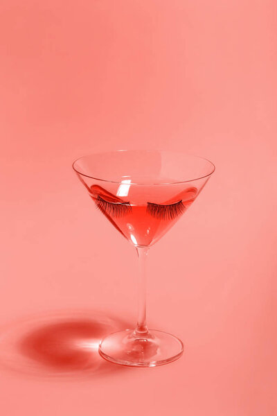 Cocktail in glass with eyelashes on living coral color backgroun