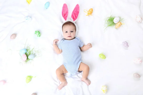 Lindo bebé con huevos de Pascua y orejas de conejo acostado en la cama blanca —  Fotos de Stock