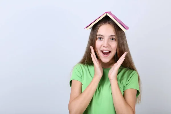 Молода дівчина тримає книгу на голові на сірому фоні — стокове фото