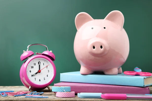 Pink piggy bank with school supplies and alarm clock on wooden t