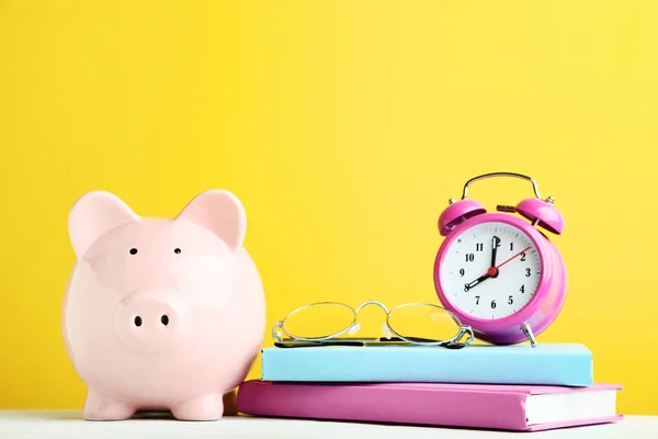 Piggy bank rosa com despertador, óculos e livros em amarelo ba — Fotografia de Stock
