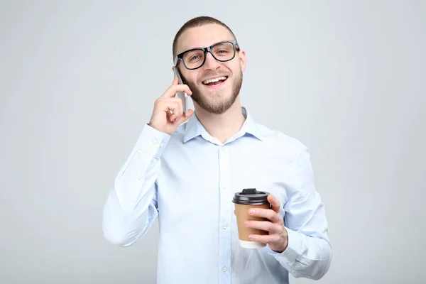 Молодий чоловік в окулярах зі смартфоном і паперовою чашкою на сірому фоні — стокове фото