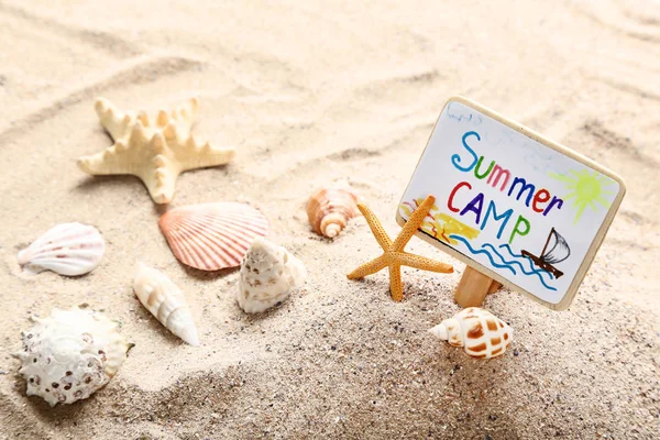 Board with text Summer camp and seashells on beach sand — Stock Photo, Image