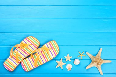 Mavi ahşap masa üzerinde deniz kabukları ile parmak arası terlik çifti