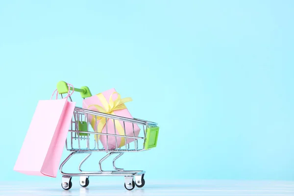 Pequeño carrito con caja de regalo y bolsa de compras sobre fondo azul —  Fotos de Stock