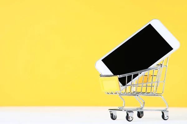 黄色の背景にスマートフォンを持つ小さなショッピングカート — ストック写真