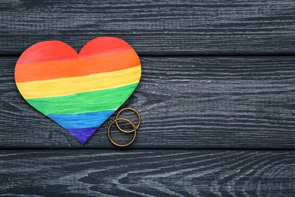 Herz aus Regenbogenpapier mit goldenen Ringen auf schwarzem Holztisch — Stockfoto