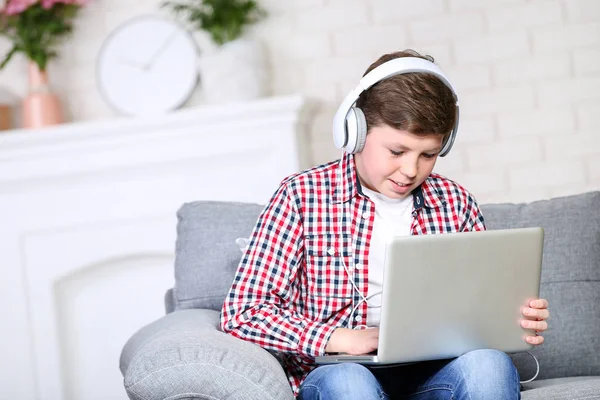 Ung dreng med laptop og hovedtelefoner sidder på sofaen - Stock-foto
