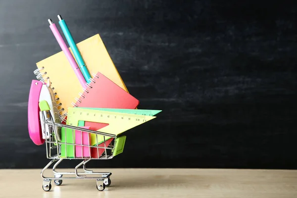 School supplies in shopping cart on blackboard background — Stock Photo, Image