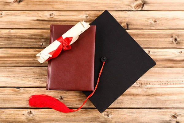 Graduation cap with diploma and book on brown wooden table — Stock Photo, Image