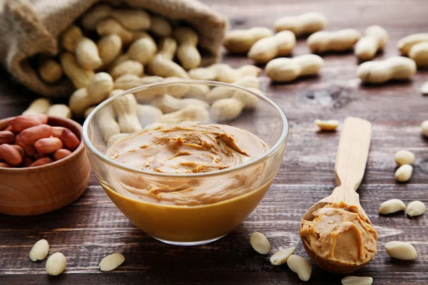 Arašídové máslo v misce a ořechy na hnědém dřevěném stole — Stock fotografie