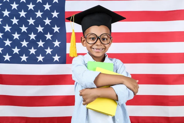 Lindo chico en gorra de graduación con libros sobre fondo de bandera americana — Foto de Stock