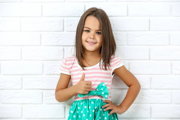 Красивая маленькая девочка показывает большой палец на белой кирпичной стене спинки — стоковое фото