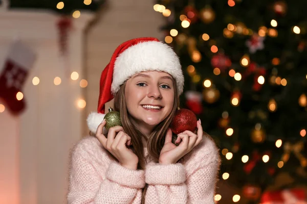 Молодая девушка в шляпе Санты с безделушками возле рождественской елки дома — стоковое фото