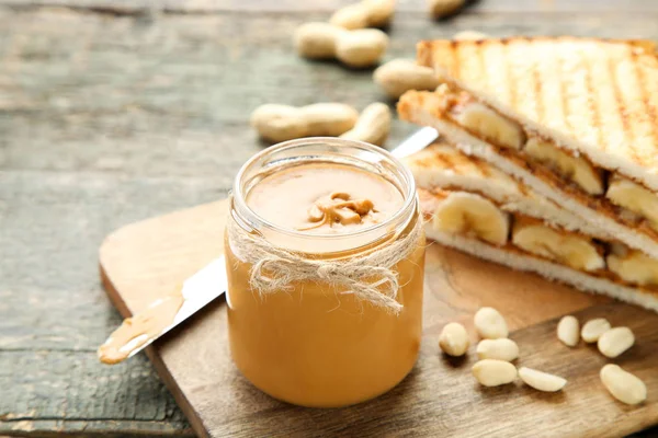 Erdnussbutter im Glas mit Brot und Nüssen auf Holztisch — Stockfoto