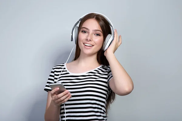 Mujer joven con auriculares y teléfono móvil sobre fondo gris — Foto de Stock