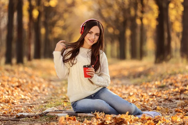 Mooie vrouw zitten met koptelefoon en papieren beker in de herfst — Stockfoto