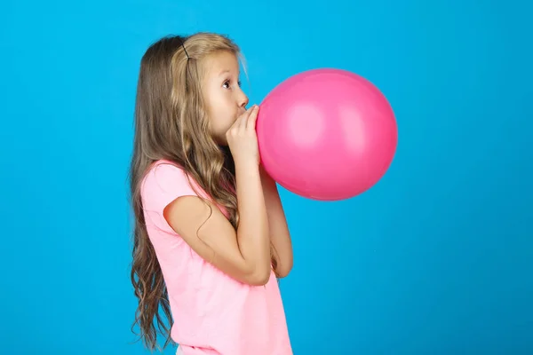 Досить маленька дівчинка дме рожева куля на синьому фоні — стокове фото