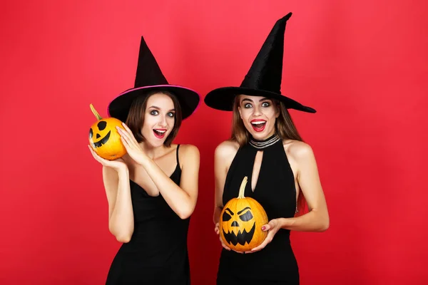 Zwei junge Frauen in schwarzen Kostümen mit Halloween-Kürbissen auf rot — Stockfoto