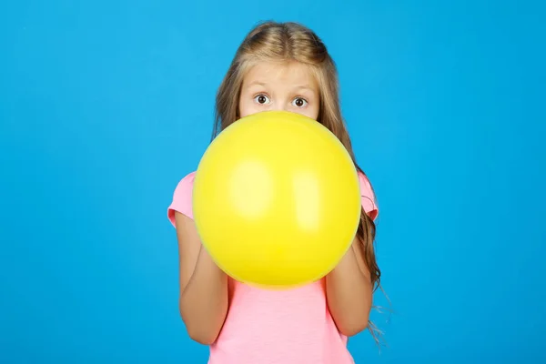 Pretty little girl blowing yellow balloon on blue background — Stock Photo, Image