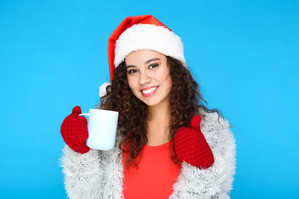Mulher americana bonita vestindo chapéu de Papai Noel e segurando xícara de chá no fundo azul — Fotografia de Stock