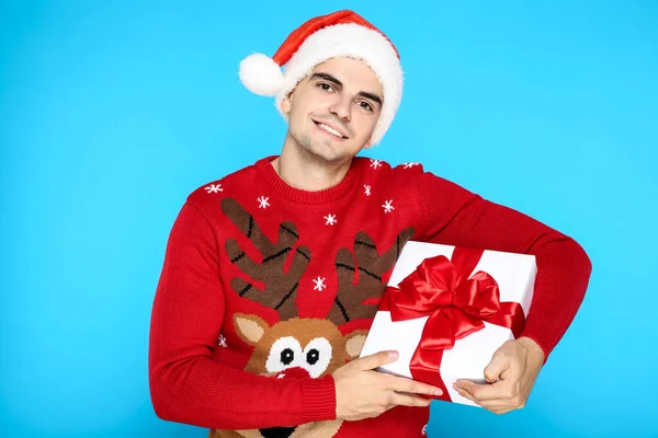 Красива людина в різдвяному светрі, капелюсі Санти і тримає подарункову коробку на синьому фоні — стокове фото