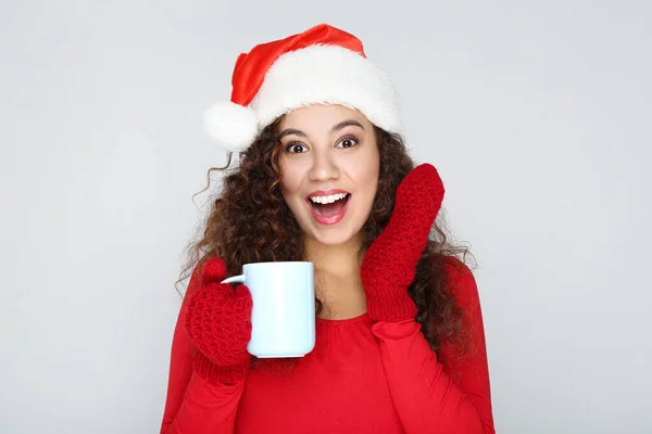Mulher americana bonita vestindo chapéu de Papai Noel e segurando xícara de chá no fundo cinza — Fotografia de Stock