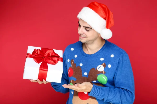 Όμορφη άνθρωπος σε χριστουγεννιάτικο πουλόβερ και Σάντα καπέλο κρατώντας κουτί δώρου σε κόκκινο φόντο — Φωτογραφία Αρχείου