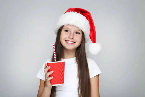 Menina no chapéu de Natal segurando copo de papel com palha no fundo cinza — Fotografia de Stock