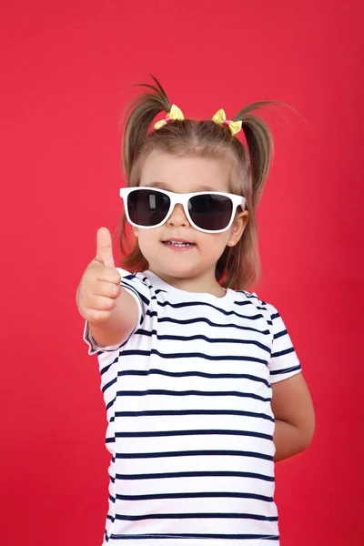 Nettes kleines Mädchen mit Sonnenbrille zeigt Daumen nach oben auf rotem Hintergrund — Stockfoto