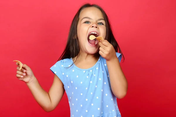 Beautiful little girl eating potato chips on red background — Zdjęcie stockowe