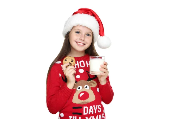Маленька дівчинка в різдвяному светрі і капелюсі тримає склянку молока і печива на білому тлі — стокове фото