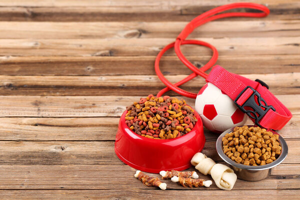 Сухой корм для домашних животных в мисках с воротником и игрушечным шариком на коричневом деревянном столе
