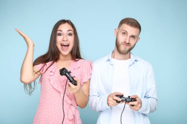 Mavi arka planda joysticklerle bilgisayar oyunu oynayan mutlu genç çift
