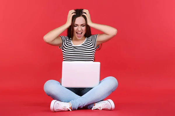 Young angry girl with laptop computer sitting on red background