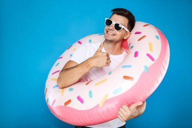 Şişme donutlu genç adam mavi arka planda baş parmağını gösteriyor.
