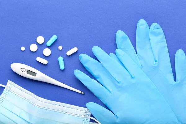 Medicine mask with pills, thermometer and pair of gloves on blue background