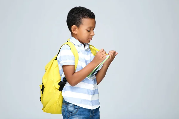 Young African American school boy writing in notepad with backpack on grey background
