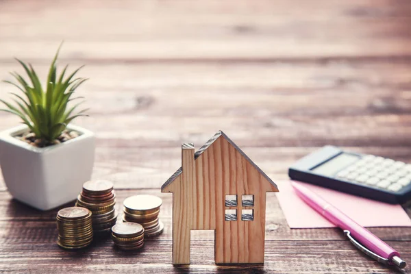 Kahverengi ahşap masa üzerinde bozuk paralar ve yeşil bitkilerle ev modeli.