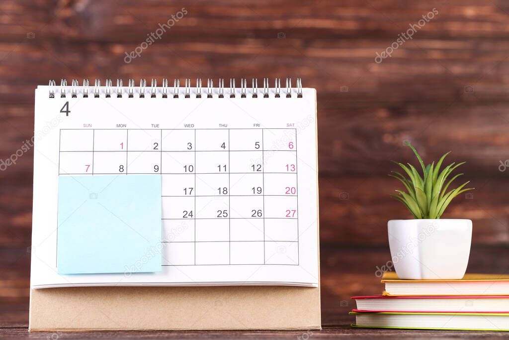 Calendar page with sticky note and green plant on brown wooden background