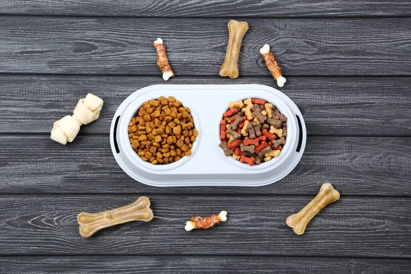 Dry pet food in bowls with bones on black wooden table