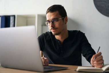 Genç adam kahve içerken masada bilgisayar, telefon ve tabletle çalışıyor.