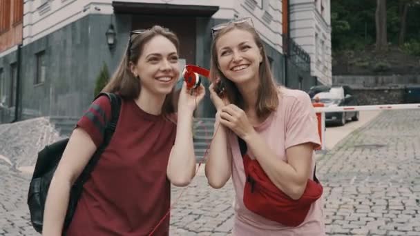 Dwie dziewczyny dzielą się czerwonymi słuchawkami, żeby posłuchać muzyki. — Wideo stockowe