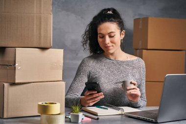 Evden bilgisayarlı ve akıllı telefonlu bir iş kadını gönderilecek ürünlerle dolu kutular hazırlıyor.