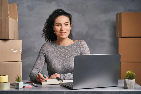 Donna d'affari che lavora da casa con laptop e blocco note, preparando scatole con prodotti per la consegna Fotografia Stock