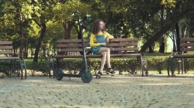 Elektrikli scooterlı kız güneşli bir sabahta parkta bankta oturuyor.