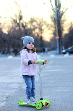 Şapkalı sevimli kız, palto ve kot pantolonla üç tekerlekli scooter'a biniyor. Akşam bahar şehir gün batımı.