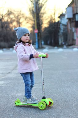 Şapkalı sevimli kız, palto ve kot pantolonla üç tekerlekli scooter'a biniyor. Akşam bahar şehir gün batımı.