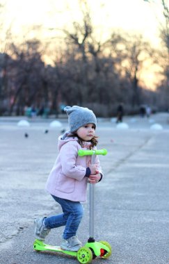Şapkalı sevimli kız, palto ve kot pantolonla üç tekerlekli scooter'a biniyor. Akşam bahar şehir gün batımı.